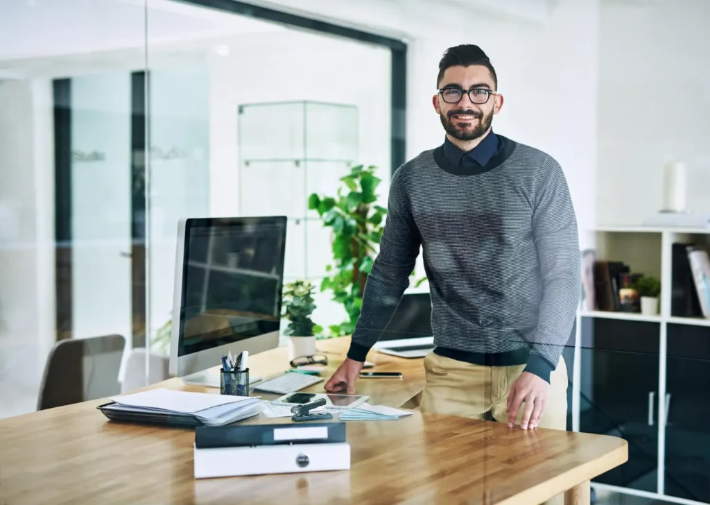 Hombre sonriente en una oficina moderna, junto a su escritorio con una computadora, representando la eficiencia en la gestión de clientes con un CRM integrado para pymes.