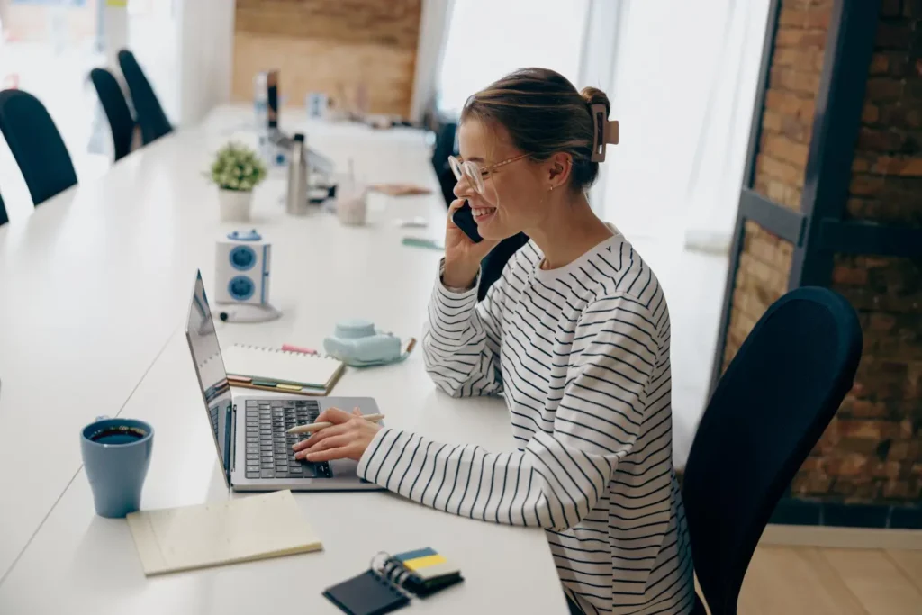 Woman discussing marketing strategy on a phone call while working on a laptop in a modern office setting.