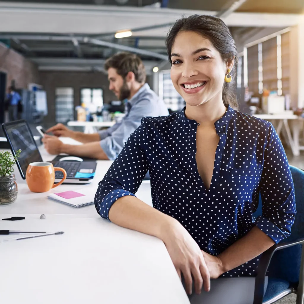 Smiling businesswoman in a polka-dot shirt sitting at a desk, working on a marketing strategy in a collaborative office environment, with a colleague in the background.