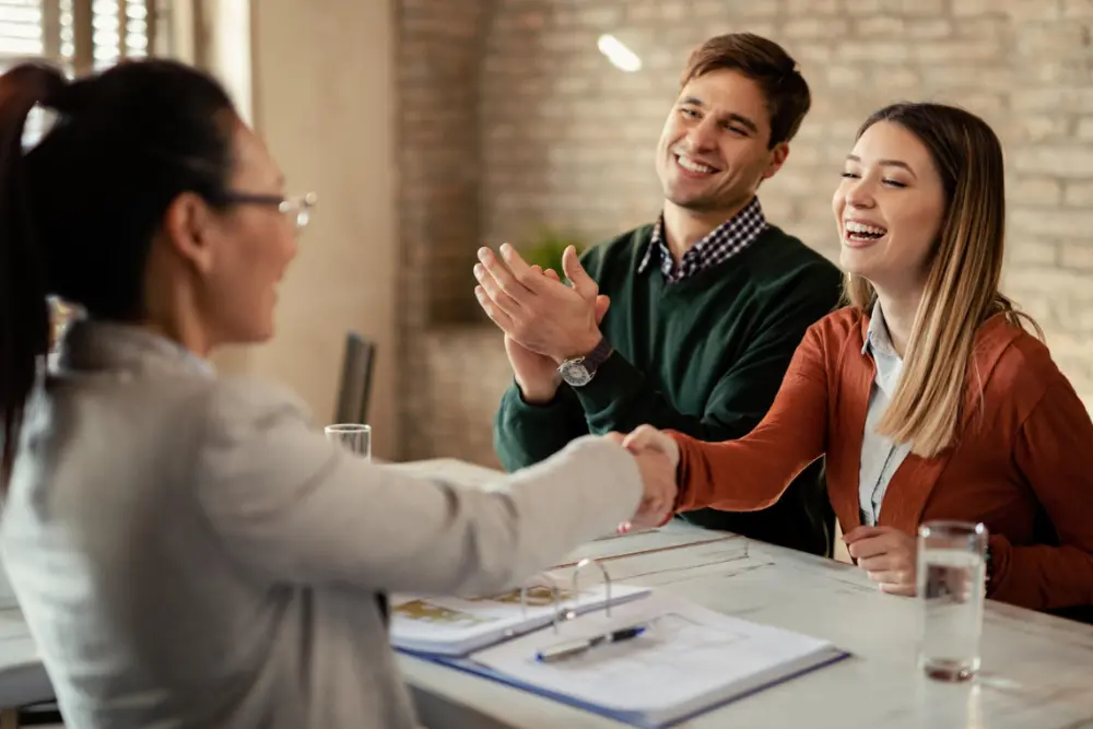 Asesor inmobiliario dando la mano a una pareja de clientes felices en una reunión, ilustrando la gestión de relaciones en un CRM inmobiliario para mejorar la satisfacción del cliente.