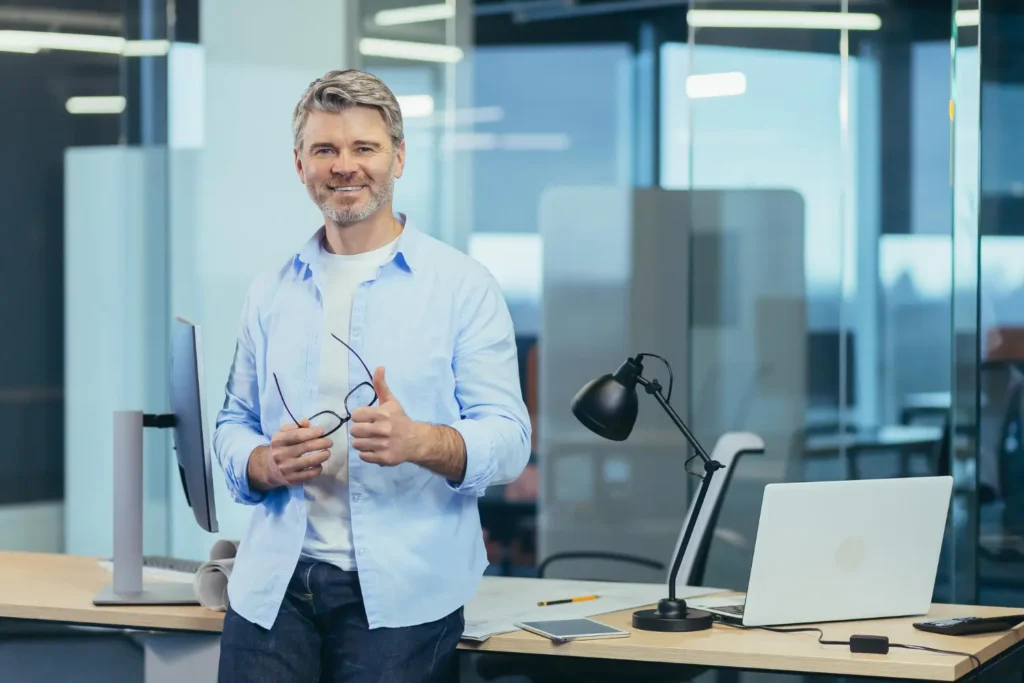 Confident business owner giving a thumbs up in a modern office, showcasing success from recent Google Ads campaigns. Desk setup with a laptop highlights digital marketing achievements.