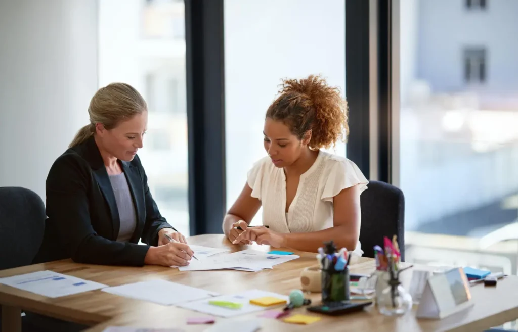 Two businesswomen sitting at a desk, analyzing documents and developing a marketing strategy in a bright office with large windows.