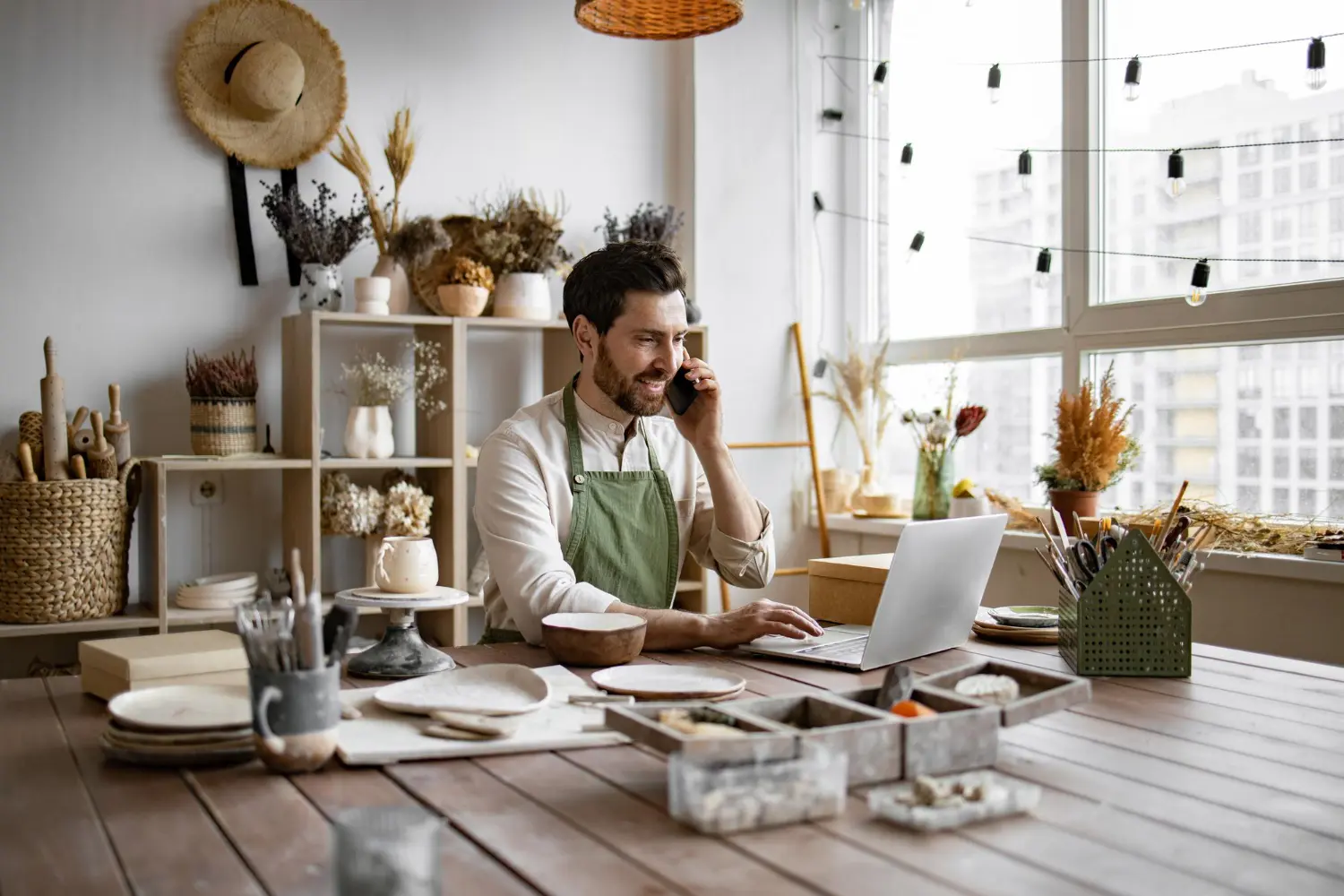 Small business owner wearing an apron, sitting at a table in a cozy home office, using a laptop and talking on the phone. The workspace is filled with pottery and crafting tools, showcasing a creative business environment. Ideal image for illustrating the use of the best online CRM for small businesses.