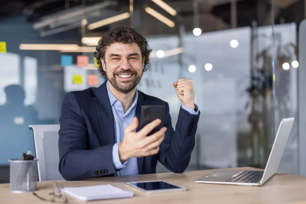 Smiling businessman in a modern office raising his fist in celebration while holding a smartphone, with a laptop and notepad on the desk, representing success in custom SEO content strategy.