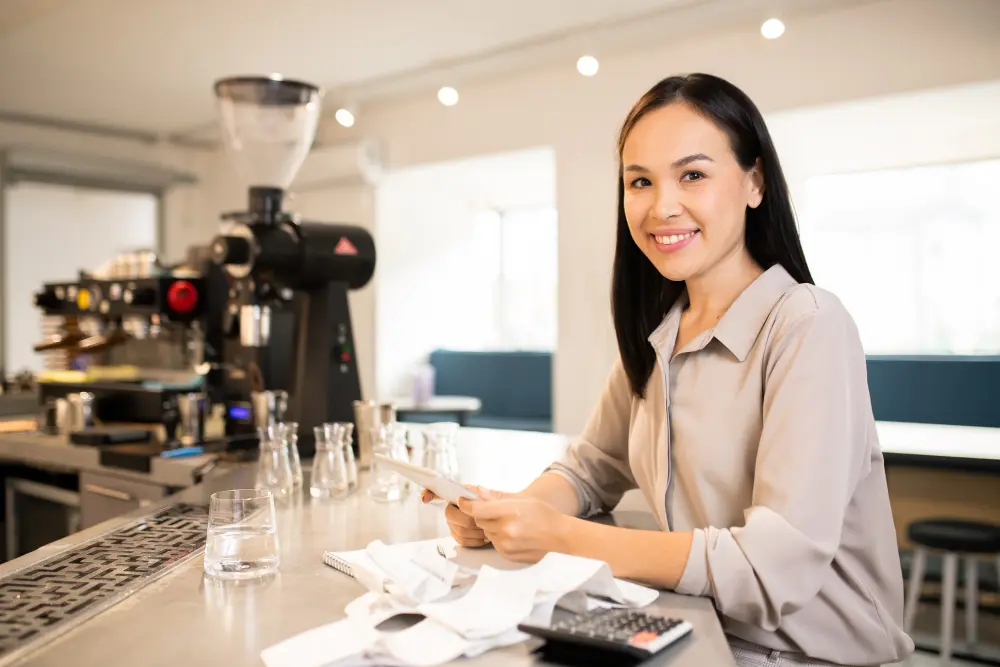 Mujer emprendedora sonriente en su cafetería revisando facturas y finanzas, representando el apoyo de una agencia de marketing para pymes en la gestión y crecimiento de pequeños negocios.