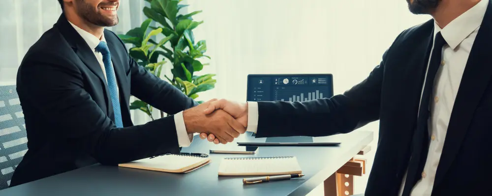 Apretón de manos entre dos ejecutivos en oficina, con laptop mostrando métricas de marketing digital